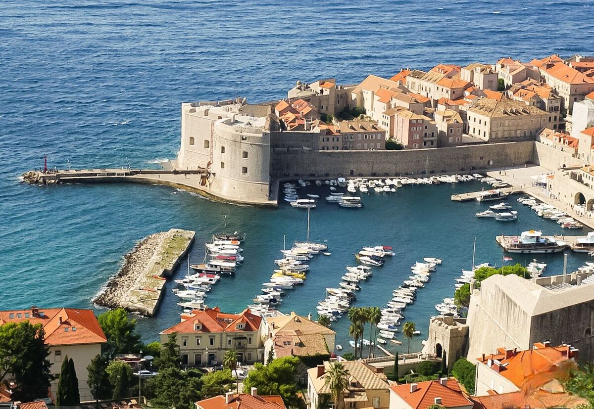 Een panoramisch uitzicht op de oude haven van Dubrovnik, met de stadsmuren en vele boten in het water. De gebouwen hebben oranje daken en de zee is helderblauw.