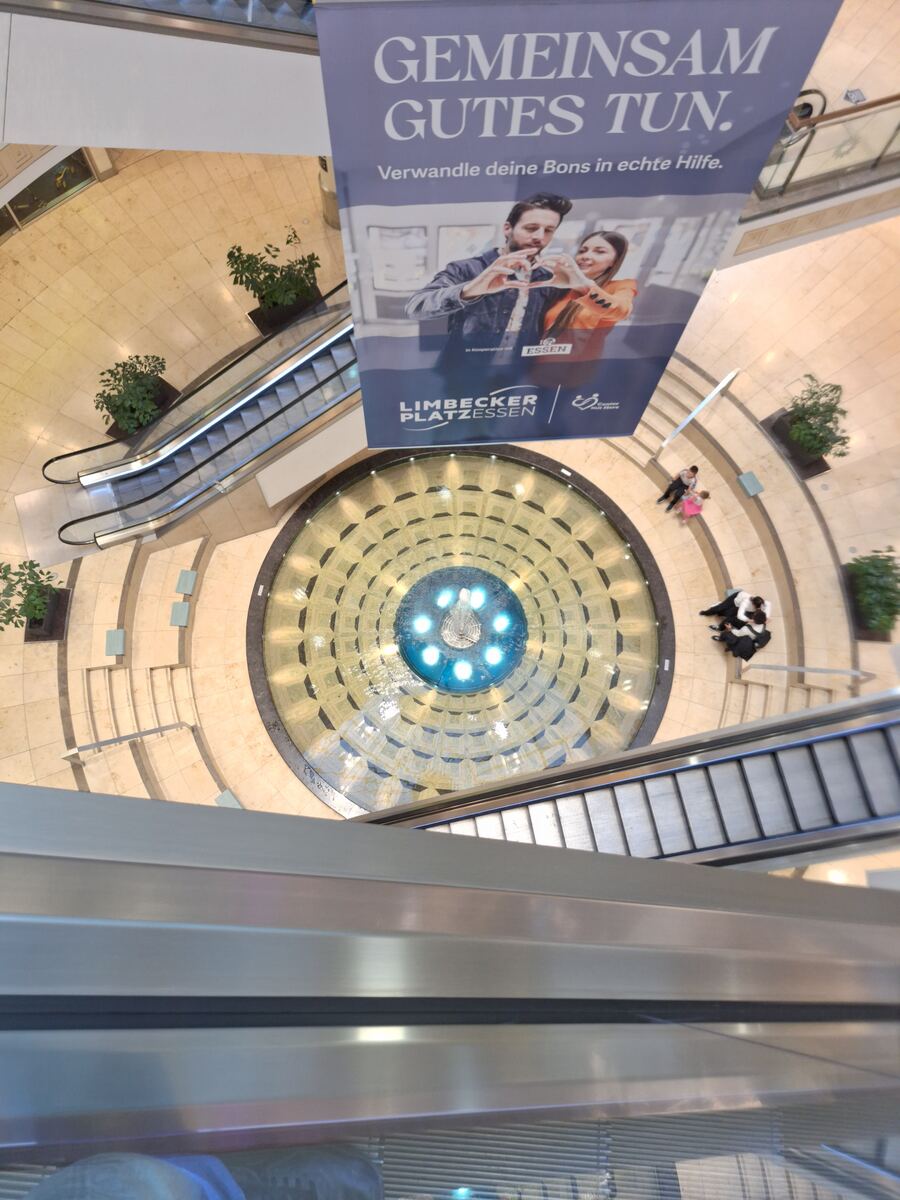 Een bovenaanzicht van een winkelcentrum met een ronde fontein in het midden, omringd door beige tegels. Er zijn roltrappen aan weerszijden van de fontein en een groot spandoek hangt boven de fontein met de tekst 'GEMEINSAM GUTES TUN.' en 'LIMBECKER PLATZESSEN'. Enkele mensen zijn zichtbaar op de benedenverdieping.