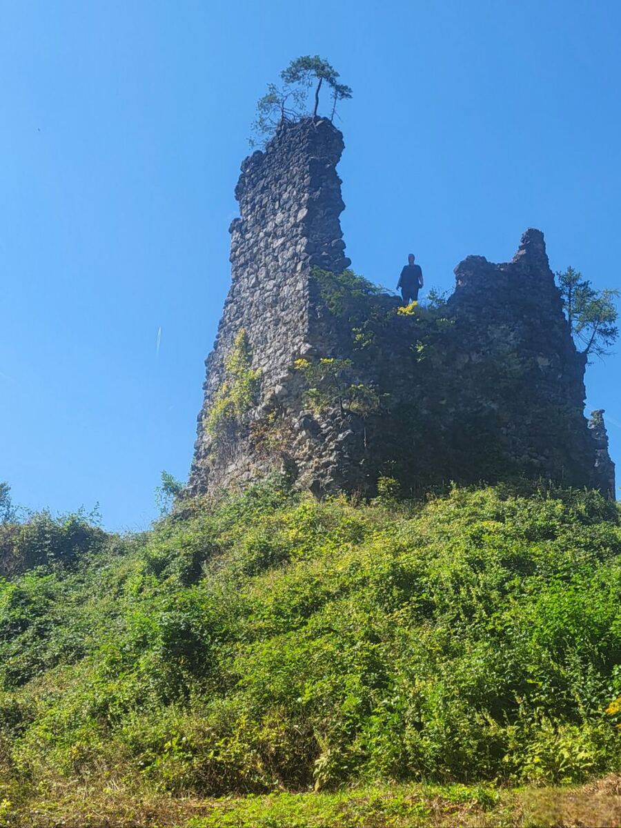 Een lage camerahoek toont de ruïne van een stenen kasteel op een met groen begroeide heuvel onder een helderblauwe lucht. Een persoon staat op een van de muren van de ruïne.