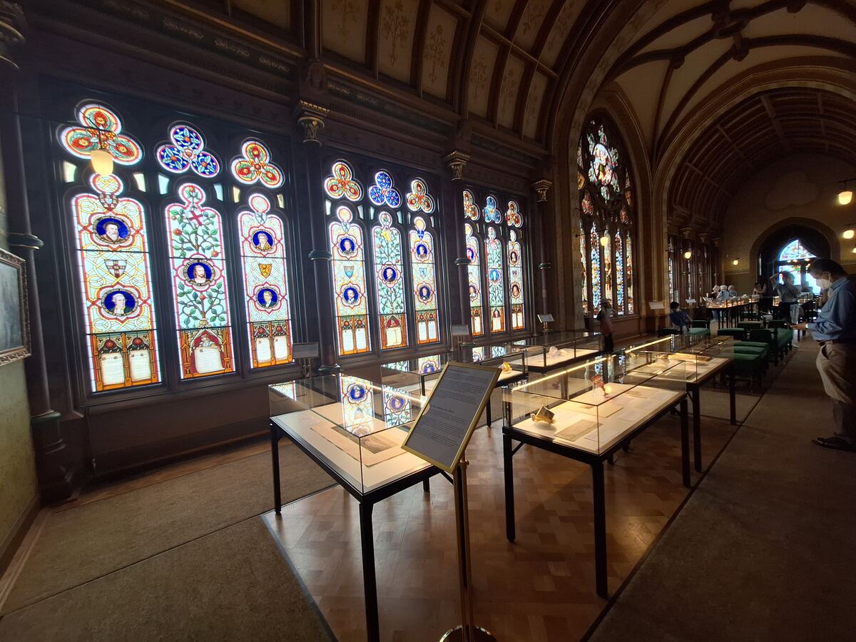Een interieur van een historisch gebouw met een lange rij gebrandschilderde ramen aan de linkerkant. Voor de ramen staan vitrines met tentoongestelde voorwerpen. De ruimte heeft een hoog, gewelfd plafond en aan de rechterkant zijn meer ramen en mensen te zien.