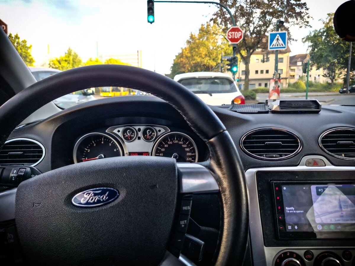 Een interieurfoto vanuit een Ford Focus met het stuur en dashboard op de voorgrond. Door de voorruit is een kruispunt te zien met een groen verkeerslicht, een stopbord en een zebrapadbord. De zon staat laag en werpt een warme gloed over de straat en de gebouwen.