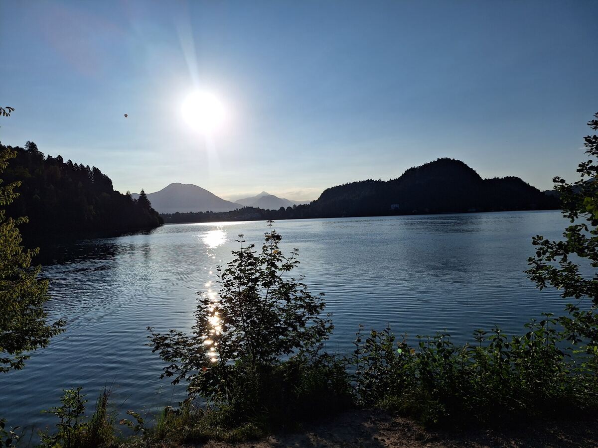 Een zonnige ochtend aan een meer met bergen op de achtergrond. De zon staat laag aan de horizon en reflecteert op het wateroppervlak. Aan de linkerkant is een hete luchtballon zichtbaar. De voorgrond bestaat uit struiken en bomen.