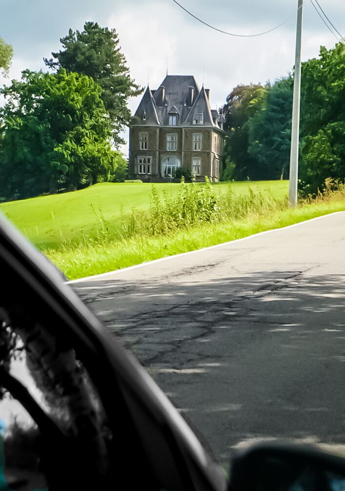 Een groot, oud kasteelachtig gebouw met meerdere torens en ramen, omringd door groen gras en bomen, gezien vanuit een rijdende auto. De weg loopt schuin omhoog en er zijn schaduwen van bomen op het asfalt zichtbaar.