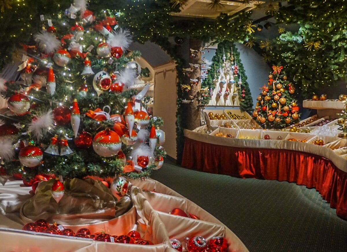Een interieurfoto van een winkel vol kerstversiering, met meerdere versierde kerstbomen en tafels vol met kerstballen en ornamenten. De kleuren rood, wit en goud overheersen. De ruimte is sfeervol verlicht met kleine lampjes.