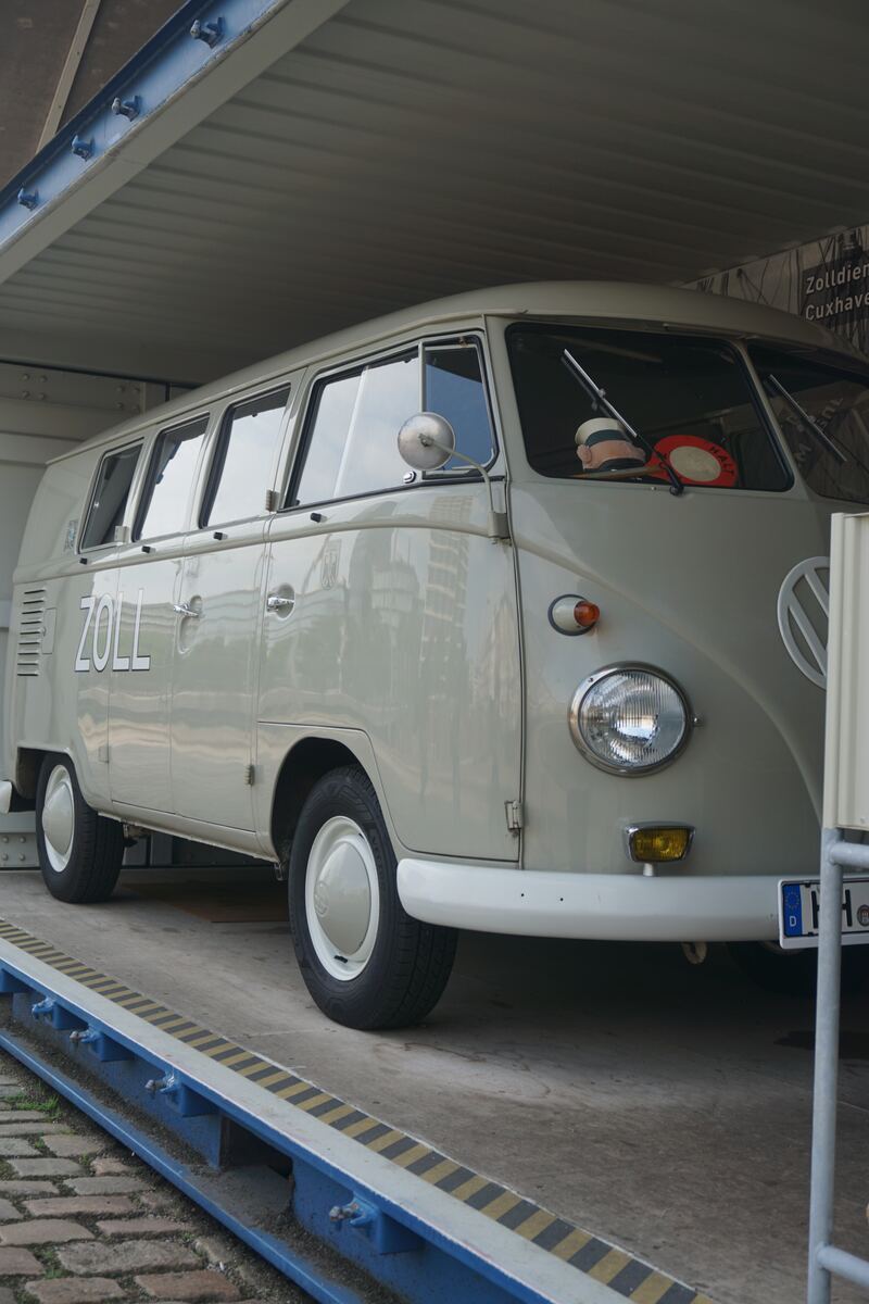 Een lichtgrijze, klassieke Volkswagen T1 'Bulli' bus staat op een verhoogd platform met geel-zwarte strepen. Op de zijkant van de bus staat in grote letters 'ZOLL' geschreven. Op het dashboard liggen een donkerblauwe pet met witte strepen en een rood 'HALT'-bordje. Boven de bus is een deel van een bord zichtbaar met de tekst 'Zolldienst Cuxhaven'. In de ramen van de bus zijn reflecties van moderne gebouwen te zien.