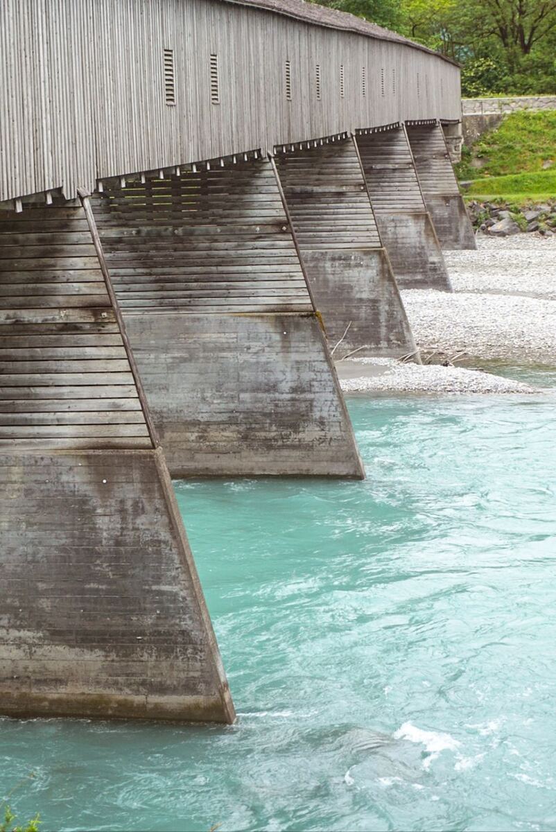 Een houten overdekte brug met meerdere betonnen pijlers die in het turquoise water van een rivier staan. De oevers zijn deels begroeid met groen en deels bedekt met kiezels.