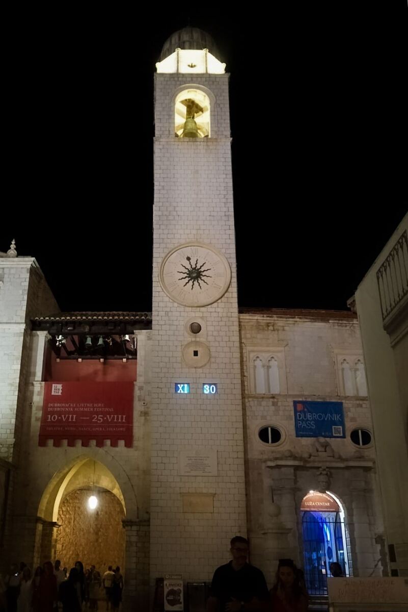 Een hoge, stenen klokkentoren met een klok en een digitaal tijdsaanduiding van 'XI 30' is 's nachts verlicht tegen een donkere hemel. Aan de linkerkant van de toren hangt een rood spandoek met tekst in het Kroatisch en Engels over het 'Dubrovnik Summer Festival'. Rechts van de toren is een gebouw met een blauw spandoek van 'Dubrovnik Pass' en een ingang met de tekst 'LUZA CENTAR'. Onder de toren zijn enkele mensen zichtbaar.