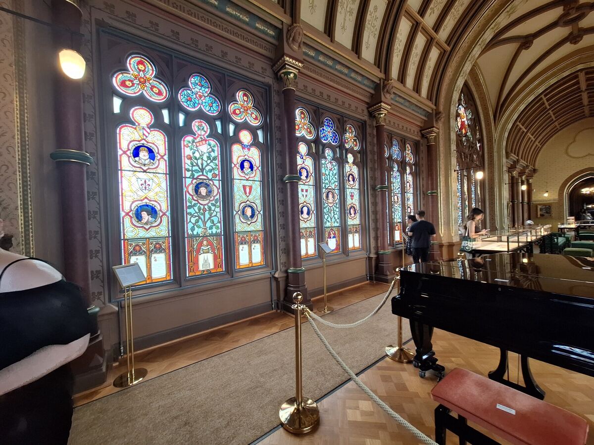 Een interieurfoto van een grote zaal met een reeks hoge, gedetailleerde glas-in-loodramen. De ramen tonen portretten en heraldische motieven in levendige kleuren. De zaal heeft een hoog gewelfd plafond met decoratieve patronen en pilaren. Op de voorgrond staat een zwarte vleugel met een rood fluwelen kruk. Er zijn enkele mensen te zien die de ramen bekijken en er staan gouden afzetpalen met touwen om een doorgang te markeren.