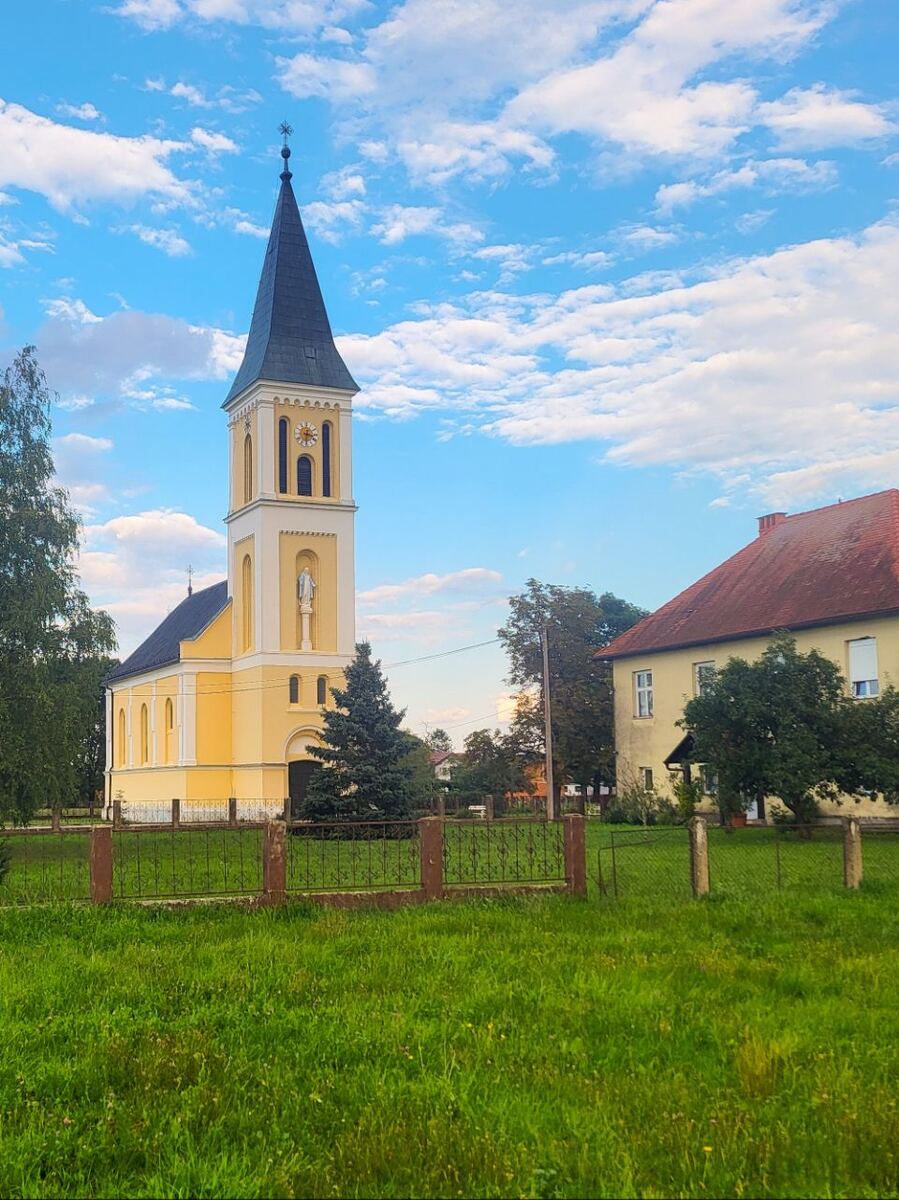 Een gele kerk met een donkergrijze spits en een klokkentoren, gelegen naast een geel woonhuis met een rood dak. Op de voorgrond is een groen grasveld met een laag hekwerk te zien. De lucht is blauw met witte wolken.