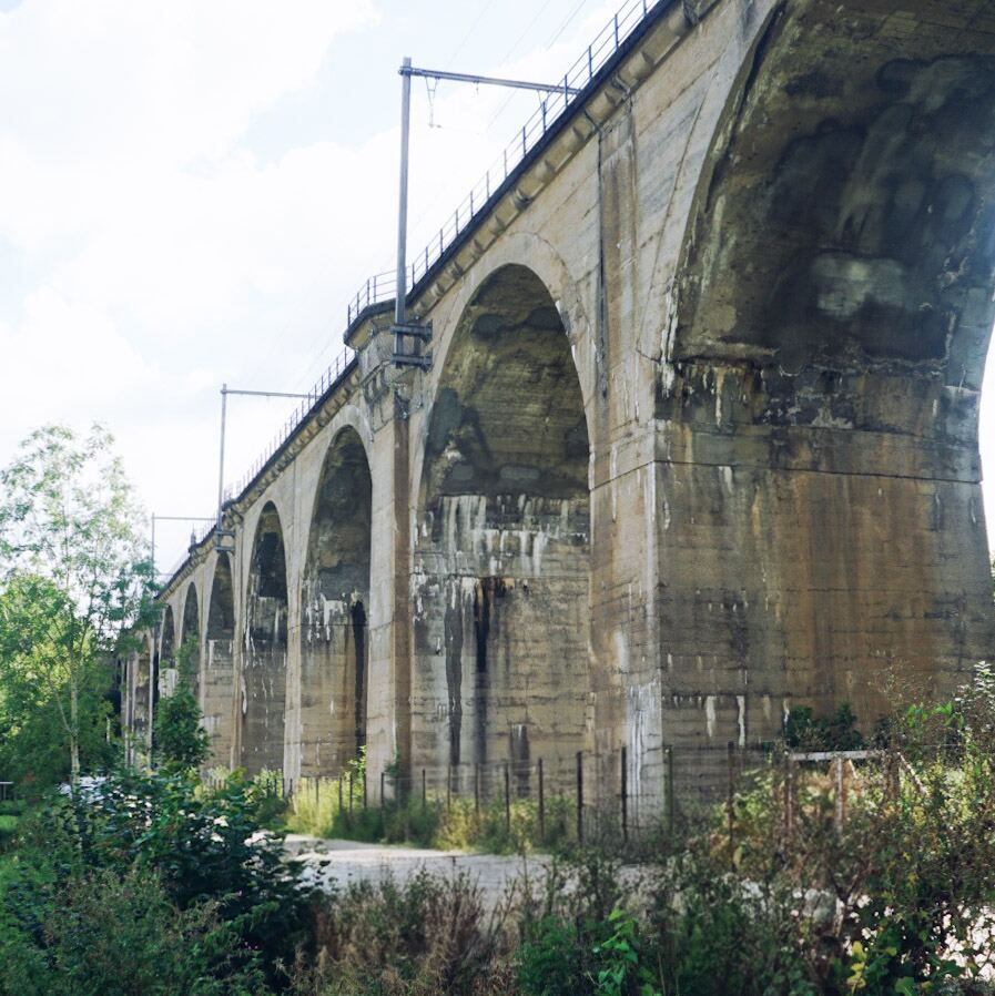 Een betonnen viaduct met meerdere bogen strekt zich uit onder een licht bewolkte hemel. De constructie is robuust en vertoont sporen van verwering en vocht. Onder het viaduct is een onverhard pad zichtbaar, omzoomd door weelderig groen en struikgewas. Bovenop het viaduct zijn spoorrails en bovenleidingen te zien.