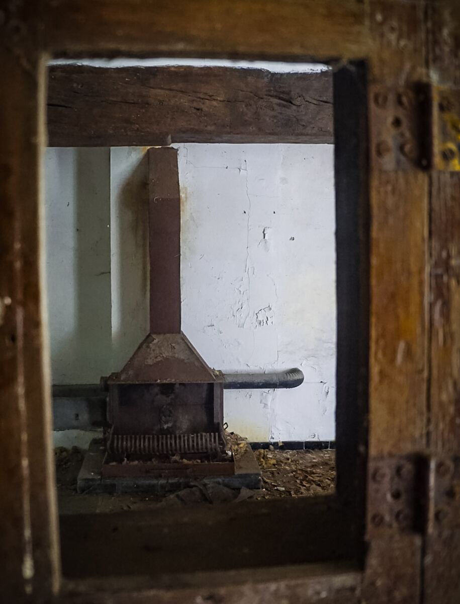 Een roestige, oude metalen haard met een schoorsteenpijp is te zien in een verlaten ruimte met afgebladderde witte muren en een houten balk bovenaan. De foto is genomen door een houten kozijn of opening.