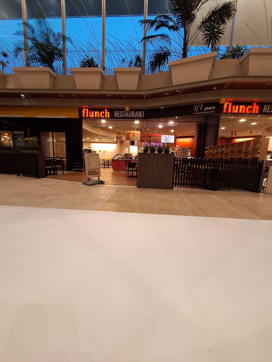 Een interieurfoto van een winkelcentrum met een restaurant genaamd 'Flunch Restaurant'. Boven het restaurant is een glazen dak zichtbaar met palmbomen en een blauwe avondlucht. Er staan tafels en stoelen voor het restaurant en er is een reclamebord met Franse tekst.