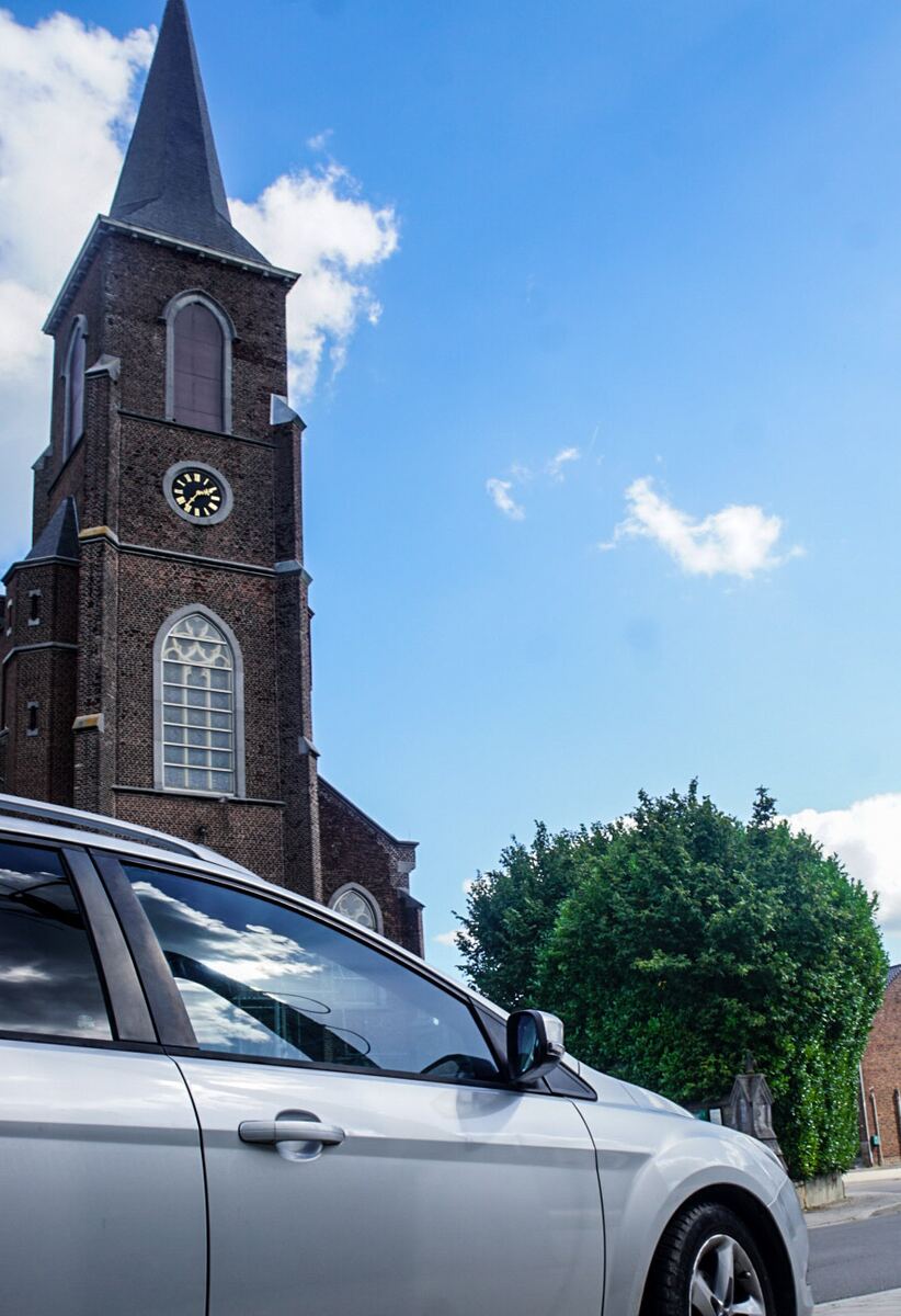 Een zilverkleurige auto staat geparkeerd voor een bakstenen kerk met een hoge spits en een klok. De lucht is blauw met enkele witte wolken. Rechts is een groene boom zichtbaar.