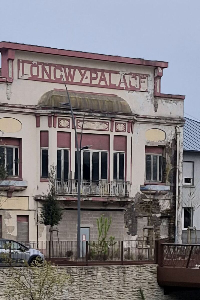 Een close-up van een oud gebouw met de tekst "LONGWY-PALACE" op de gevel. Het gebouw heeft een balkon met een metalen reling en ramen met rolluiken. De gevel toont tekenen van verval met afgebladderde verf en beschadigd stucwerk. Een lantaarnpaal staat voor het gebouw en op de achtergrond is een deel van een ander gebouw met een schuin dak zichtbaar. Een auto is deels zichtbaar aan de linkerkant van de foto, achter een hek.