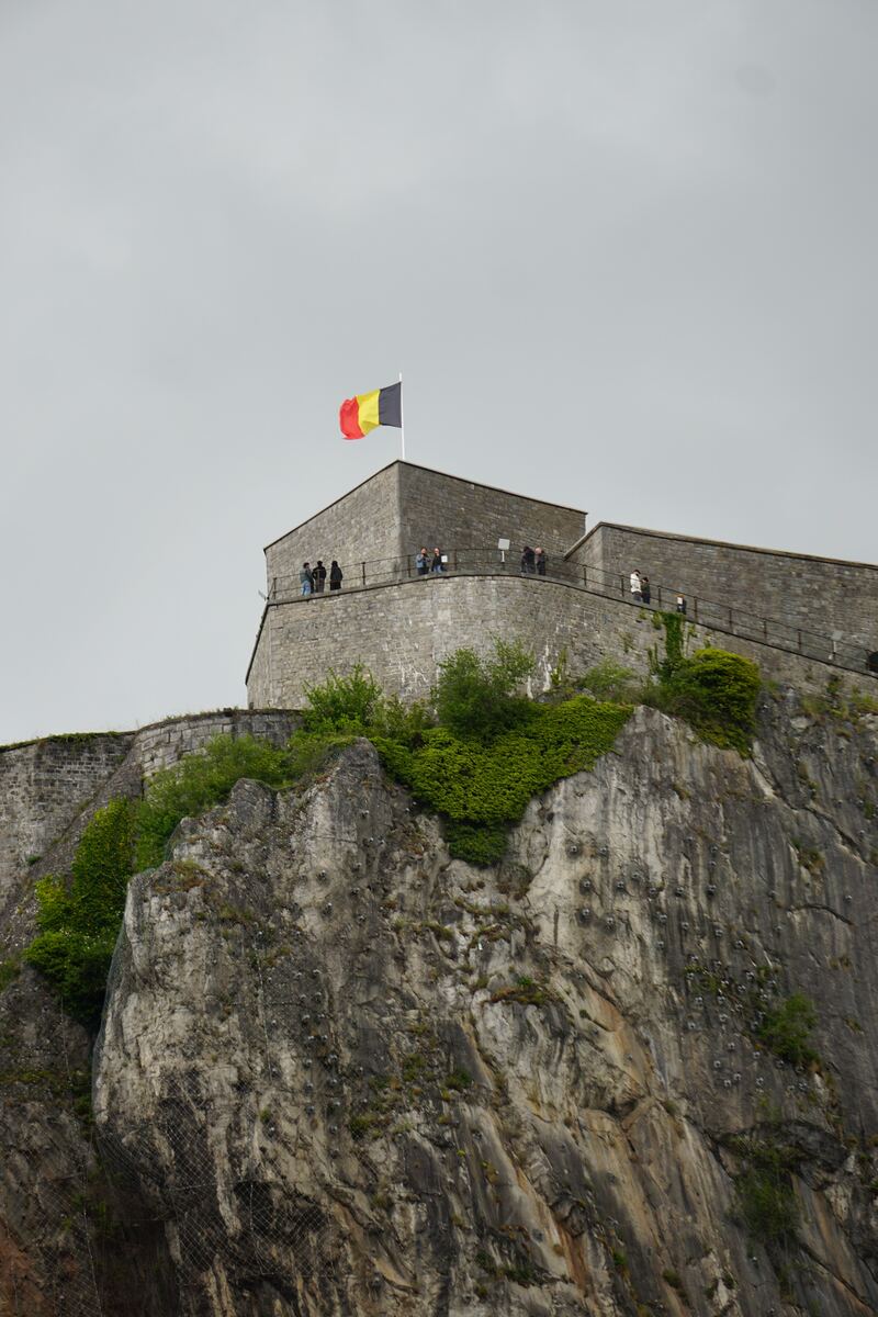 Een Belgische vlag wappert bovenop een stenen vestingmuur, die deel uitmaakt van een citadel op een steile rotswand. Er zijn mensen zichtbaar op de vestingmuur en de rotswand is deels bedekt met groen en verstevigd met netten.