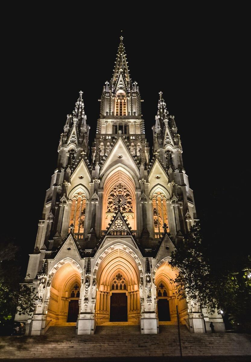 Een hoge, gotische kerk met meerdere torens en spitsen, sfeervol verlicht in de avond. De gevel toont gedetailleerde architectuur met roosvensters en spitsbogen. De verlichting accentueert de details en geeft de kerk een warme gloed tegen de donkere nachtelijke hemel.