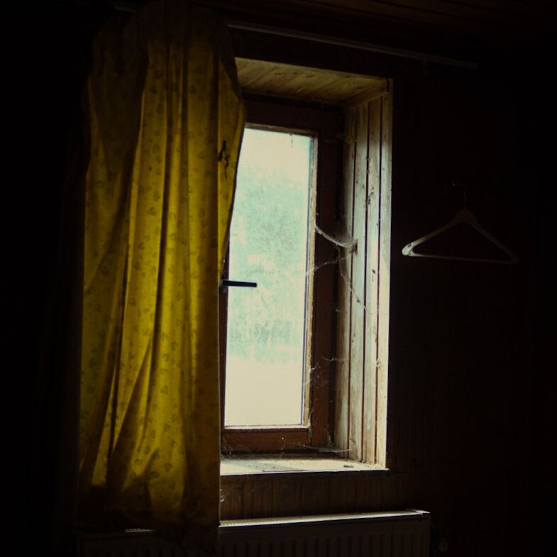 Een donkere kamer met een raam waar licht doorheen valt. Een geel gordijn hangt voor een deel van het raam. Het kozijn is van hout en er zijn spinnenwebben zichtbaar. Rechts van het raam hangt een kledinghanger aan de muur. Onder het raam is een radiator te zien.