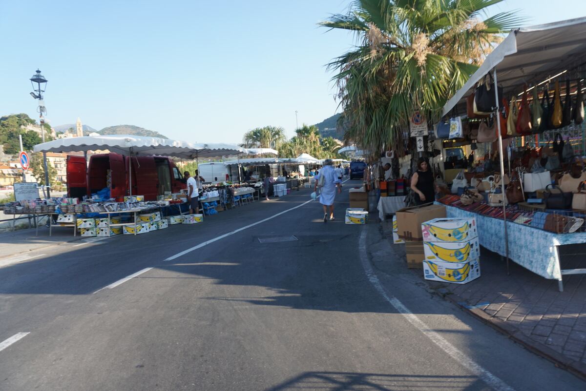 Een openluchtmarkt langs een weg met diverse kraampjes onder witte parasols. Er zijn verkopers en enkele bezoekers zichtbaar. Aan de kraampjes hangen tassen en liggen andere koopwaar. Aan de linkerzijde staat een rode bestelbus en verderop een witte. Palmboomen staan langs de rechterkant van de weg, met heuvels en gebouwen in de verte onder een helderblauwe lucht.