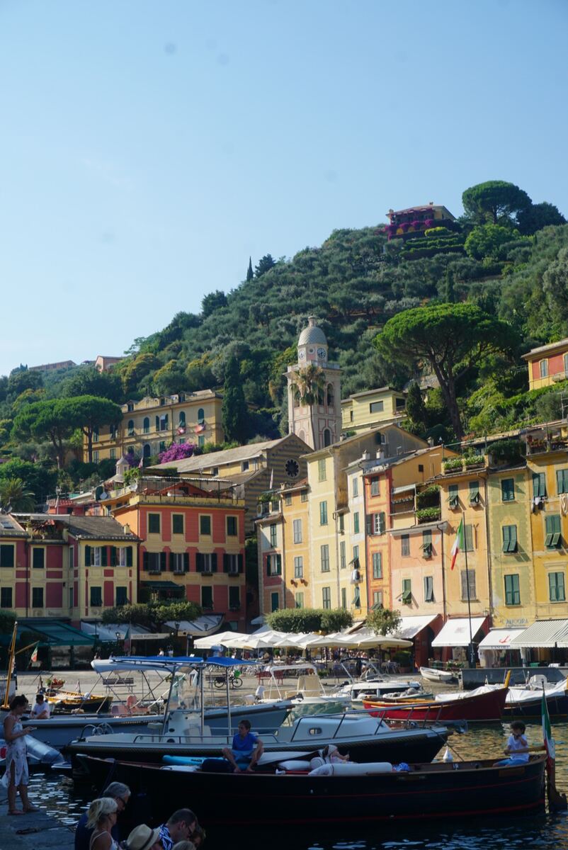 Een zonnige middag in een levendige Italiaanse havenplaats, waar kleurrijke gebouwen op een steile, beboste heuvel liggen. Een klokkentoren met een koepel is prominent zichtbaar tussen de huizen. In de voorgrond liggen diverse boten in het water, met mensen op de kade en in de boten. Witte parasols en buiten zitjes van eetgelegenheden zijn te zien langs de waterkant.