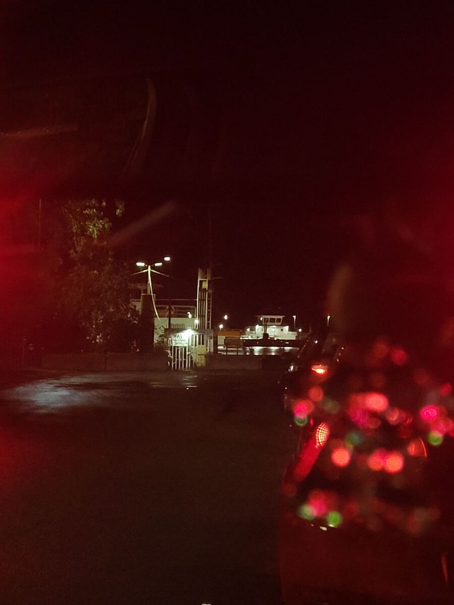 Een nachtelijke opname vanuit een auto, gericht op een veerbootterminal. We zien de achterkant van een rode auto met bokeh-effecten van de achterlichten op de voorgrond. In de verte zijn de verlichte structuren van een veerbootterminal zichtbaar, met een veerboot aangemeerd of in de buurt. De omgeving is donker, met enkele straatverlichting en reflecties op het natte wegdek.