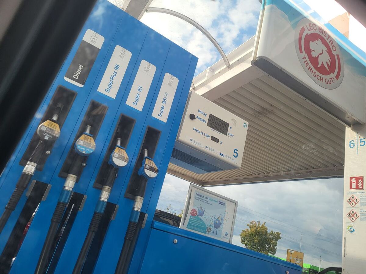 Een close-up van een blauwe brandstofpomp met verschillende soorten brandstof, waaronder Diesel, SuperPlus 98, Super 95 en Super 95 E10. Boven de pomp hangt een display met de getankte hoeveelheid en prijs. Op de achtergrond is een deel van een tankstationoverkapping te zien met een logo van 'LEO BISTRO TIERISCH GUT!' en een bewolkte lucht.