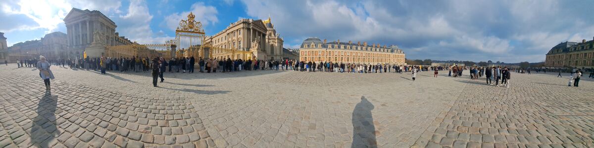 Een panoramische foto van de Cour d'Honneur van het Paleis van Versailles, met een grote groep mensen die in de rij staan voor de gouden hekken. De zon schijnt fel en er zijn enkele wolken aan de blauwe lucht. De gebouwen van het paleis zijn duidelijk zichtbaar, met hun klassieke architectuur en gouden accenten.