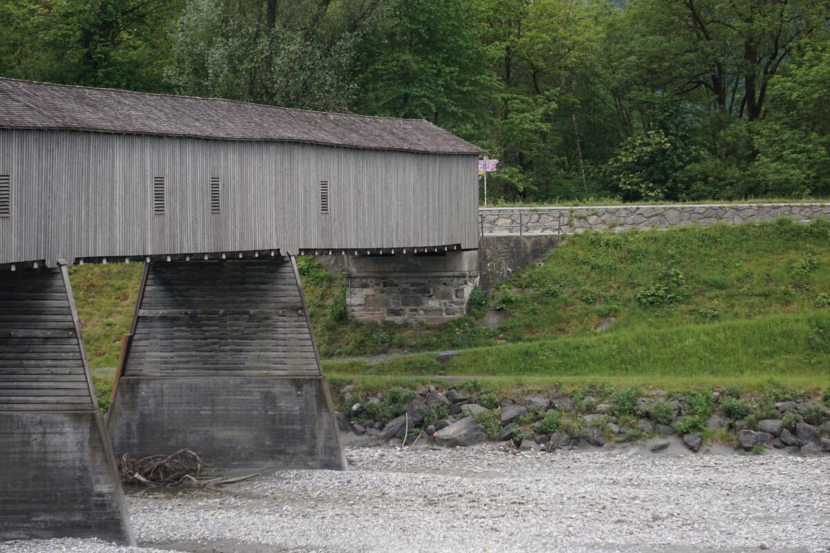 Een houten overdekte brug met een pannendak overspant een droge rivierbedding met kiezelstenen. De brug heeft verticale houten planken en enkele smalle openingen. Aan de rechterkant van de brug loopt een weg met een stenen muur en een metalen reling, omgeven door groen gras en bomen. Een klein, meerkleurig verkeersbord is zichtbaar achter de brug.