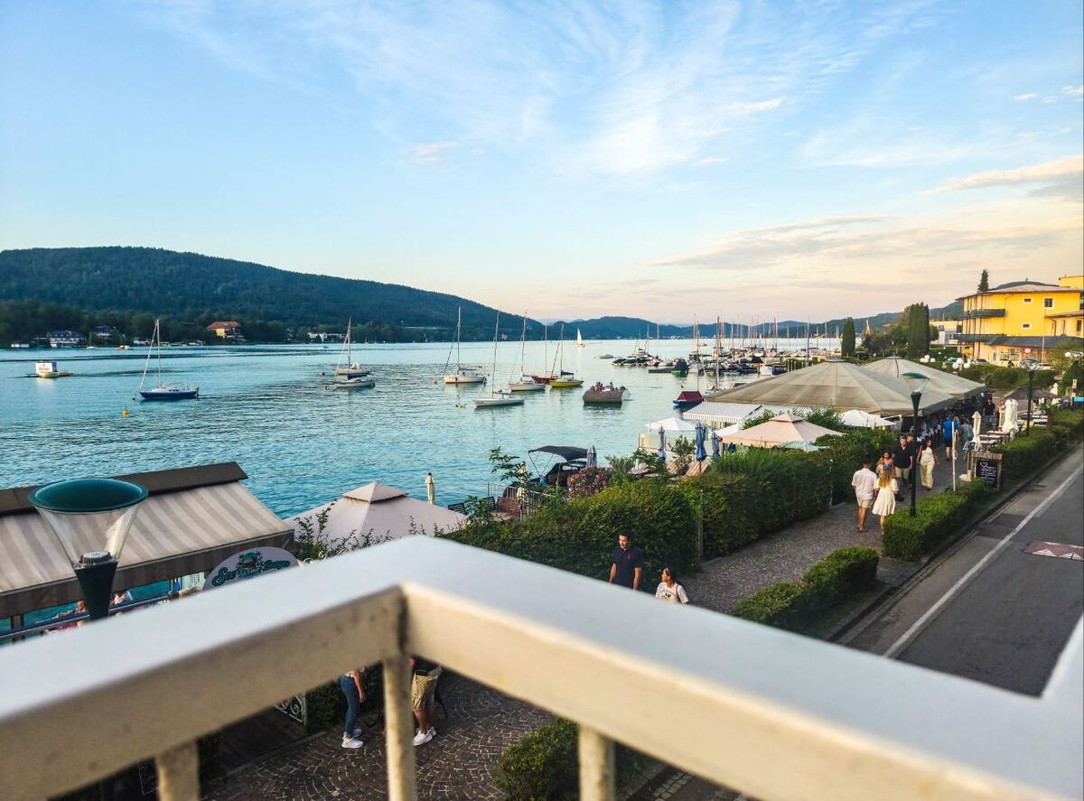 Een avondgezicht op een meer met zeilboten en een boulevard langs de oever. Op de boulevard lopen mensen en zijn er terrassen met parasols. Op de achtergrond zijn beboste heuvels te zien en de lucht is licht bewolkt met een zonsonderganggloed.
