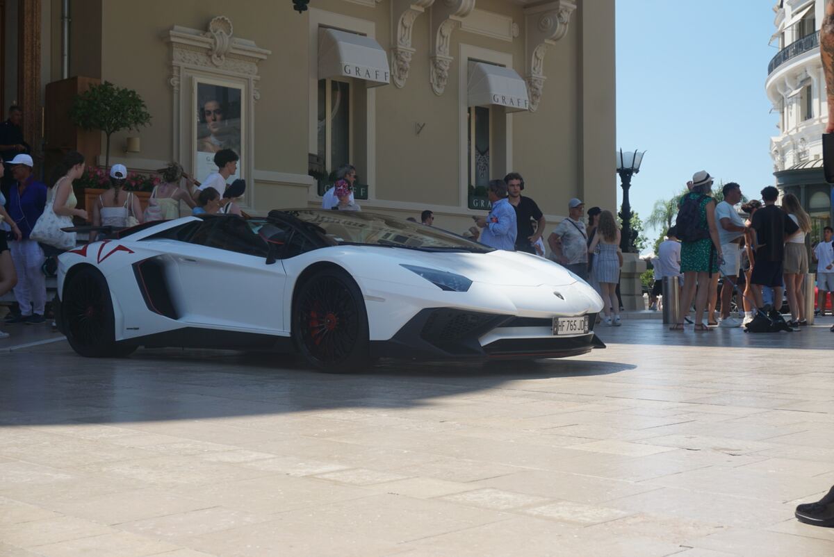Een witte Lamborghini Aventador convertible staat geparkeerd op een geplaveid plein voor een gebouw met luxe winkels. Op de achtergrond zijn diverse mensen te zien die rondkijken en wandelen. De zon schijnt helder op de scène.