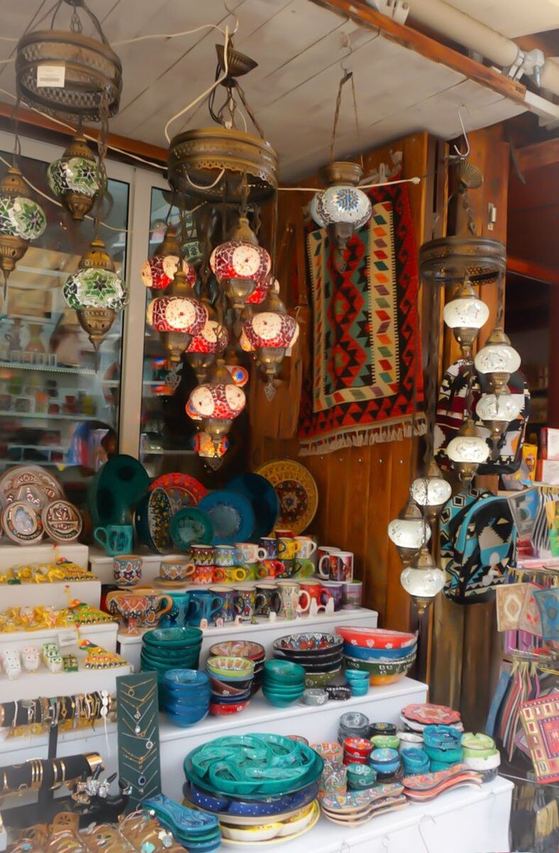 Een kraam vol met kleurrijke souvenirs, waaronder mozaïeklampen, keramische schalen, kopjes, borden, en sieraden. Op de achtergrond hangt een traditioneel geweven tapijt.