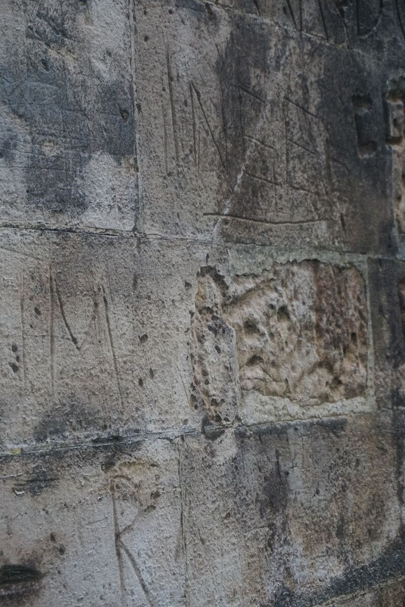 Een close-up van een oude, grijze stenen muur met zichtbare voegen en verschillende ingekraste letters en symbolen. De steen heeft een ruwe textuur en vertoont tekenen van verwering en donkere verkleuringen op sommige plekken. Een van de stenen in het midden is duidelijk beschadigd en heeft een onregelmatige vorm.