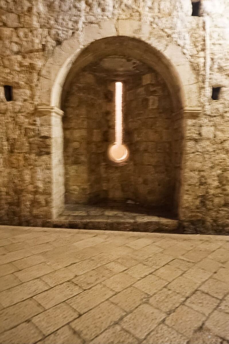 Een verticale foto van een stenen muur met een boogvormige nis en een smalle, verticale opening die naar beneden toe breder wordt en een ronde vorm aanneemt. Er schijnt licht door deze opening. De muur is opgebouwd uit onregelmatige stenen en de grond is bedekt met geplaveide stenen. De foto is 's nachts genomen en de omgeving is spaarzaam verlicht.