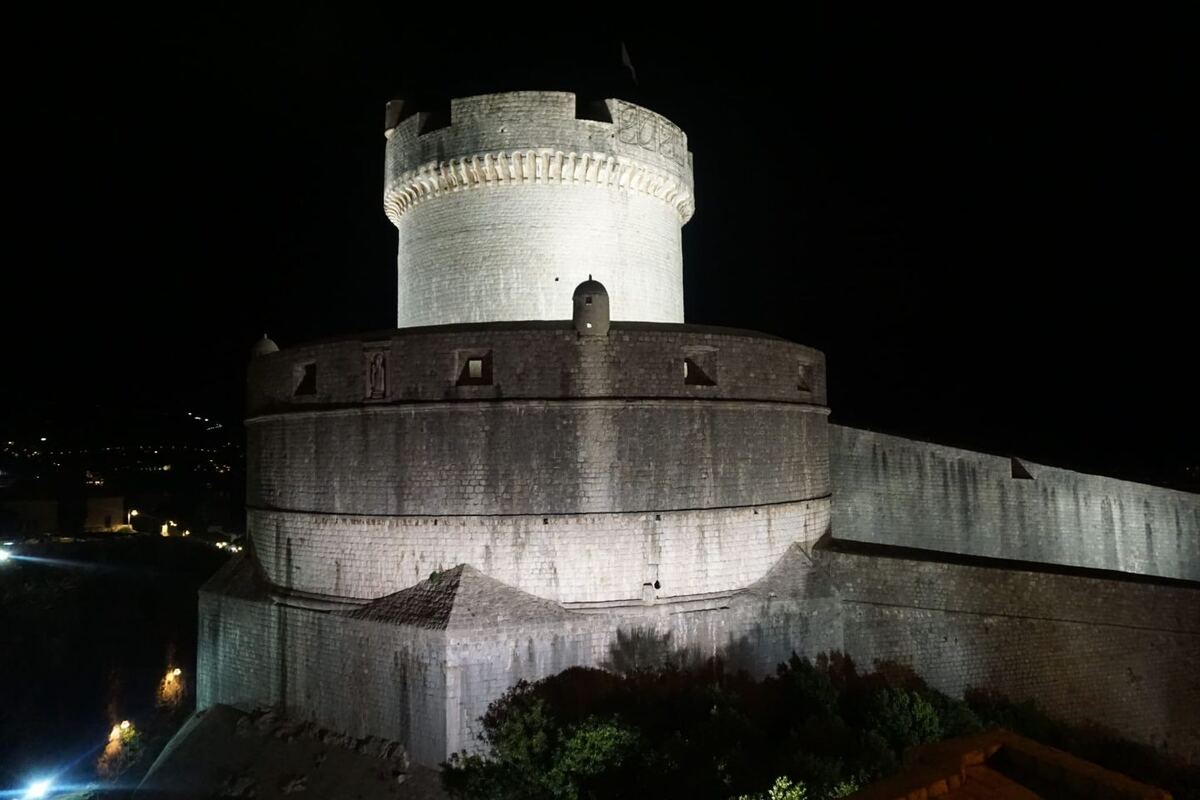 Een ronde stenen toren met kantelen, verlicht in het donker, staat naast een lange stenen muur. Op de achtergrond zijn de lichtjes van een stad te zien.