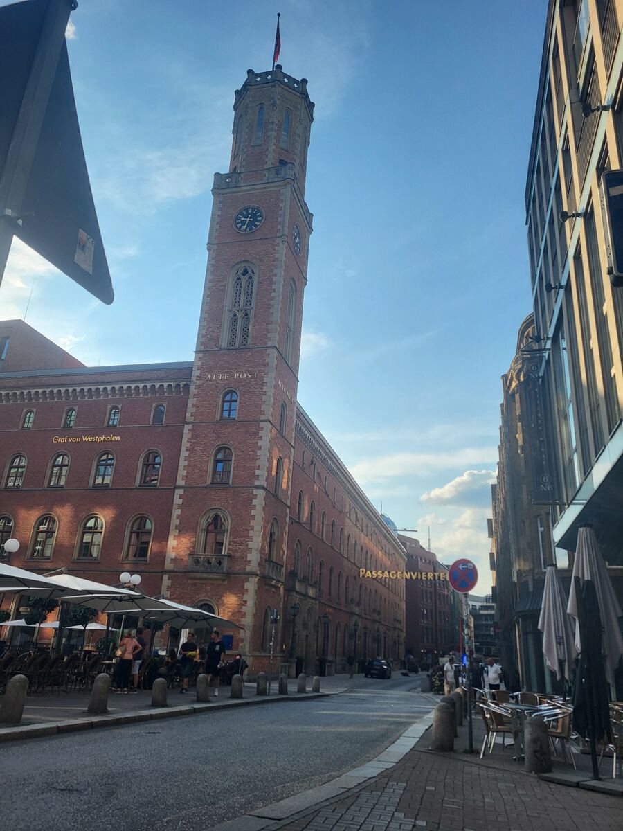 Een hoge, bakstenen toren met een klok en een vlag bovenaan, onderdeel van een groter gebouw met de tekst 'ALTE POST' en 'Graf von Westphalen'. Aan de overkant van de straat is een moderner gebouw met de tekst 'PASSAGENVIERTEL' te zien. Er staan tafels en stoelen van terrassen langs de stoep en enkele mensen zijn zichtbaar.