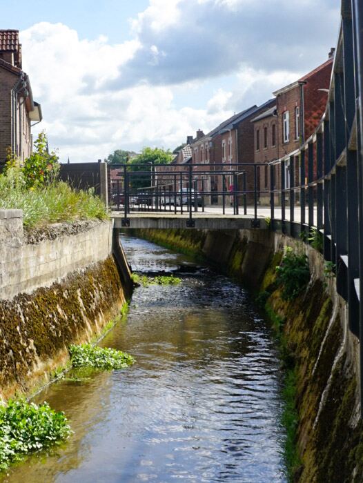 Een waterloop stroomt onder een kleine brug door, omringd door bakstenen huizen en begroeide oevers onder een bewolkte lucht.