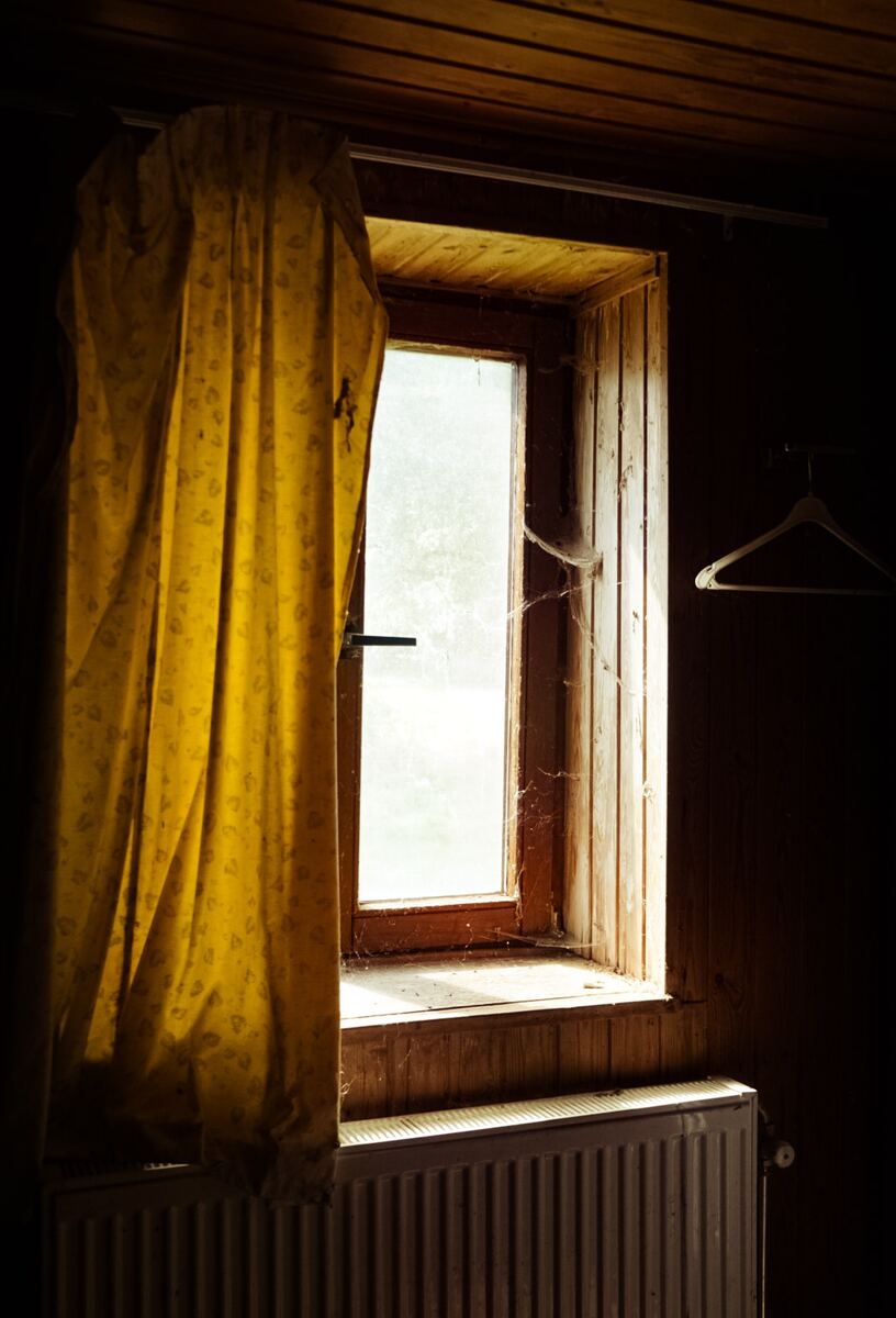 Een donkere kamer met een houten raamkozijn waar zonlicht doorheen valt. Een geel gordijn hangt aan de linkerkant. Op het raamkozijn en in de hoeken zijn spinnenwebben zichtbaar. Onder het raam staat een witte radiator en aan de rechterkant hangt een kledinghanger aan de houten wand.