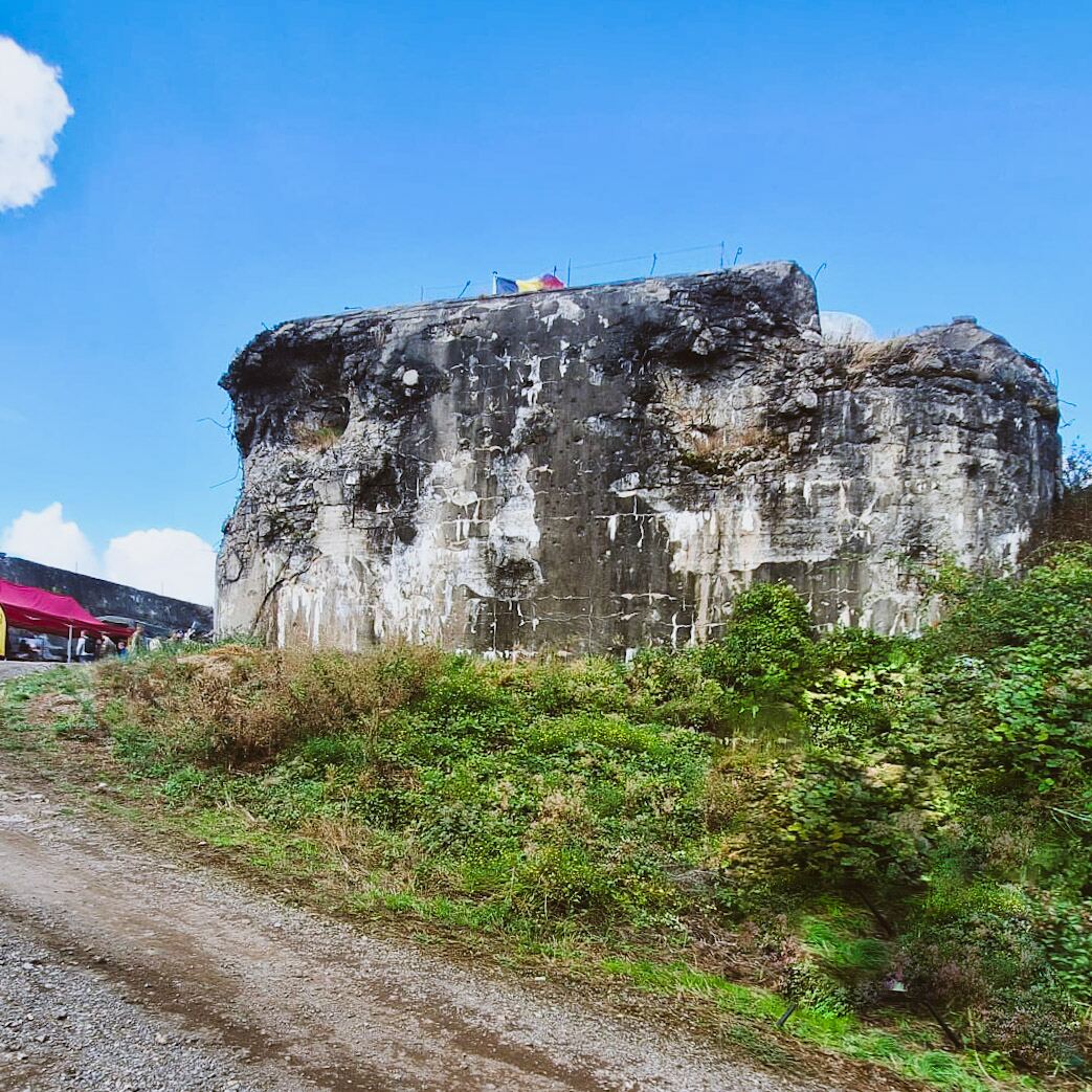 Een grote, verweerde betonnen bunker staat onder een helderblauwe lucht met enkele witte wolken. De bunker is deels begroeid met groen en heeft aan de bovenzijde enkele vlaggen. Op de voorgrond is een onverhard pad te zien en linksachter staat een rode tent met mensen eromheen.
