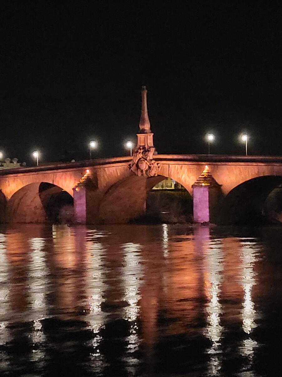 Een stenen boogbrug is 's avonds verlicht met warme oranje en paarse accenten, weerspiegelend in het donkere water eronder. Op de brug staan lantaarnpalen en in het midden bevindt zich een opvallende obelisk-achtige decoratie met beeldhouwwerk. De lucht is volledig donker.
