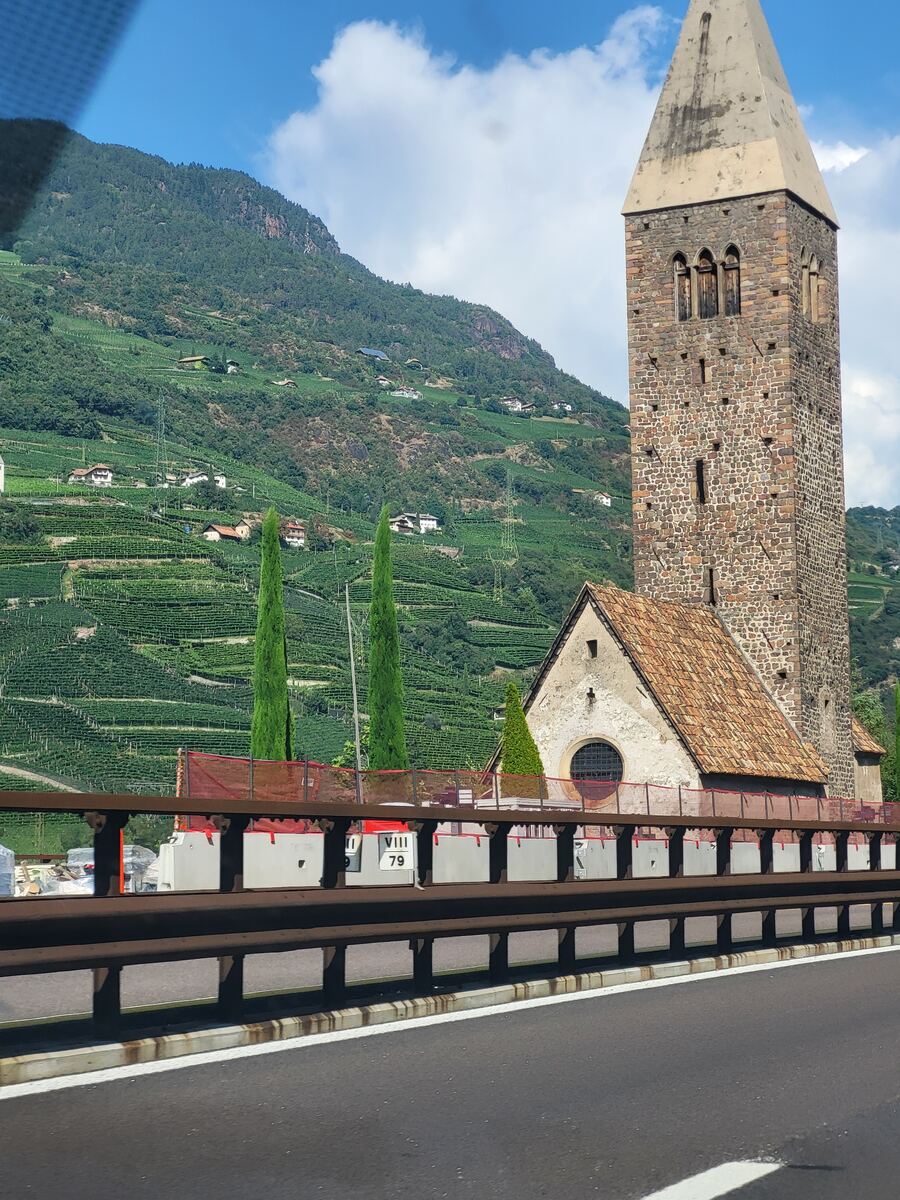 Een foto genomen vanuit een rijdende auto, met op de voorgrond een vangrail en een deel van de weg. Achter de vangrail is een stenen kerktoren met een spitse top zichtbaar, naast een kleiner kerkgebouw met een pannendak. Op de achtergrond is een steile berghelling te zien die volledig bedekt is met wijngaarden en verspreide huizen. De lucht is blauw met enkele witte wolken.