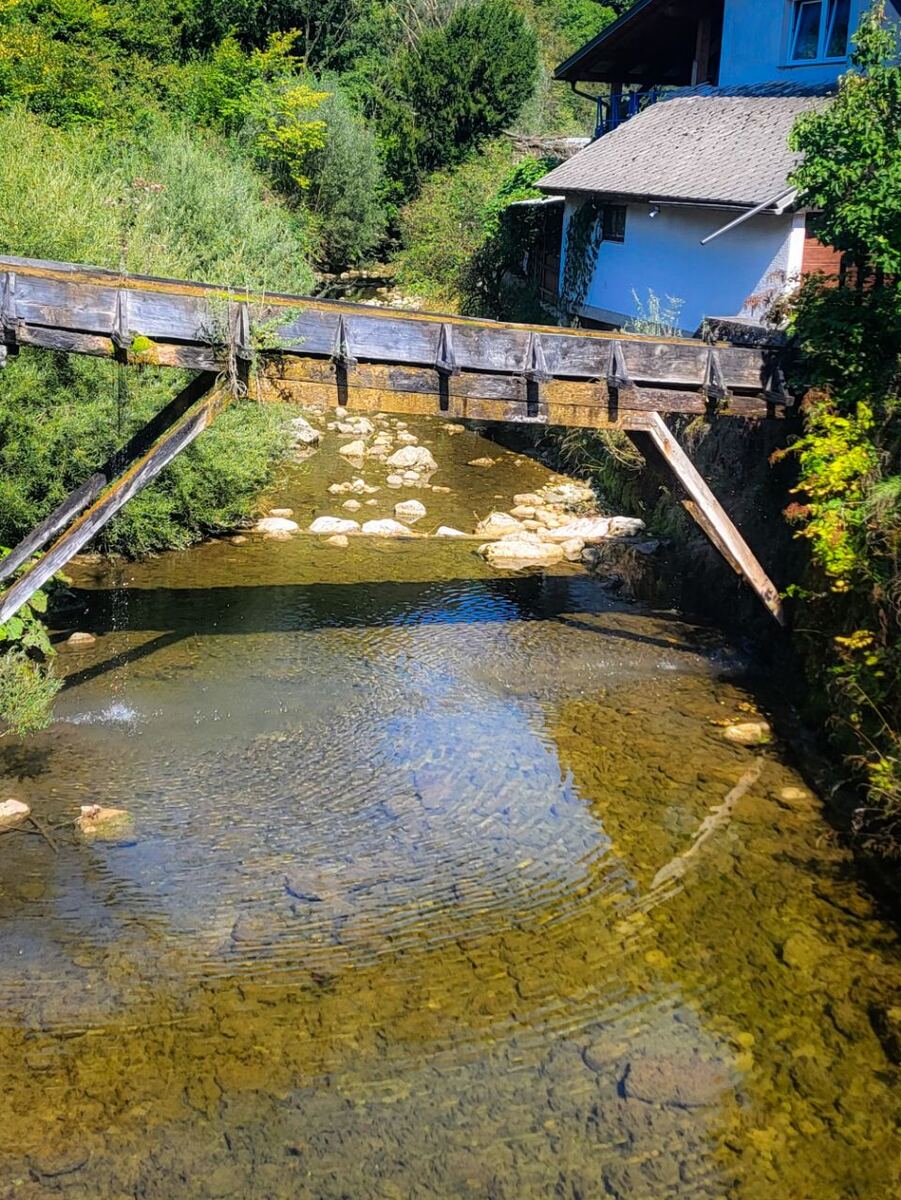Een houten brug over een ondiepe beek met veel stenen op de bodem. De beek stroomt door een groene omgeving met bomen en struiken. Op de rechteroever is een wit huis met een grijs dak zichtbaar, deels begroeid met klimop. De zon schijnt en het wateroppervlak reflecteert de lucht.