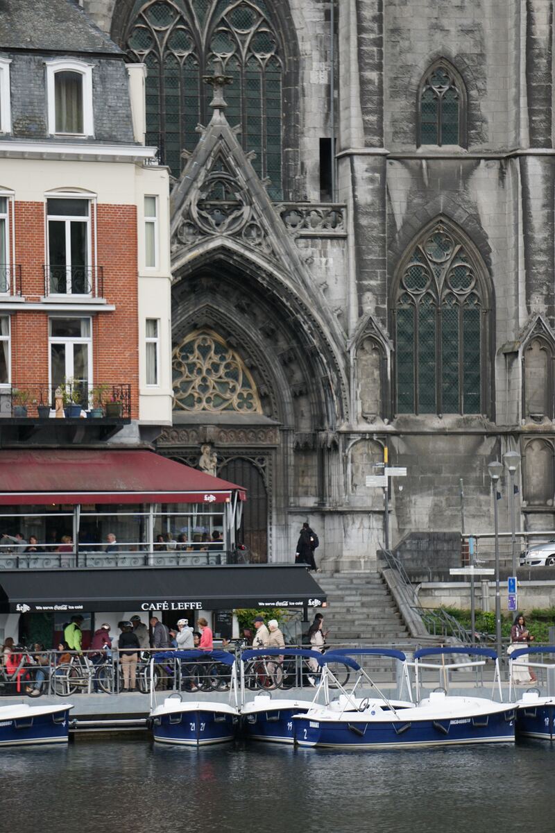 Een close-up van de Maas in Dinant, met op de voorgrond blauw-witte plezierbootjes. Daarachter een kade met mensen, een terras van 'Café Leffe' en een rood bakstenen gebouw. Op de achtergrond is de gotische architectuur van een grote kerk te zien, met gedetailleerde ramen en bogen.