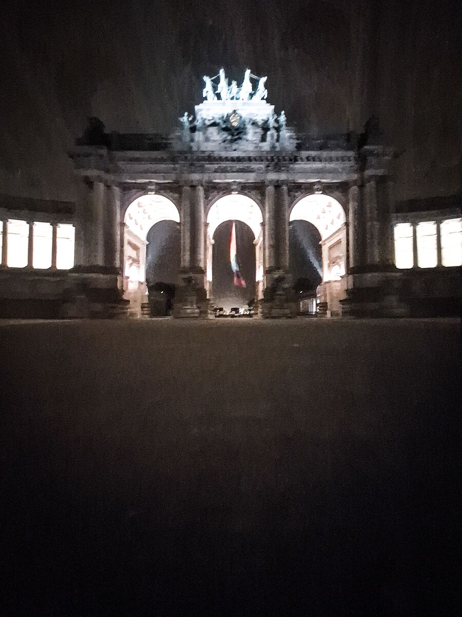 Een nachtelijke foto van de triomfboog van het Jubelpark in Brussel, verlicht met een Belgische vlag die in de middelste boog hangt. De lucht is donker en de voorgrond is een leeg plein.