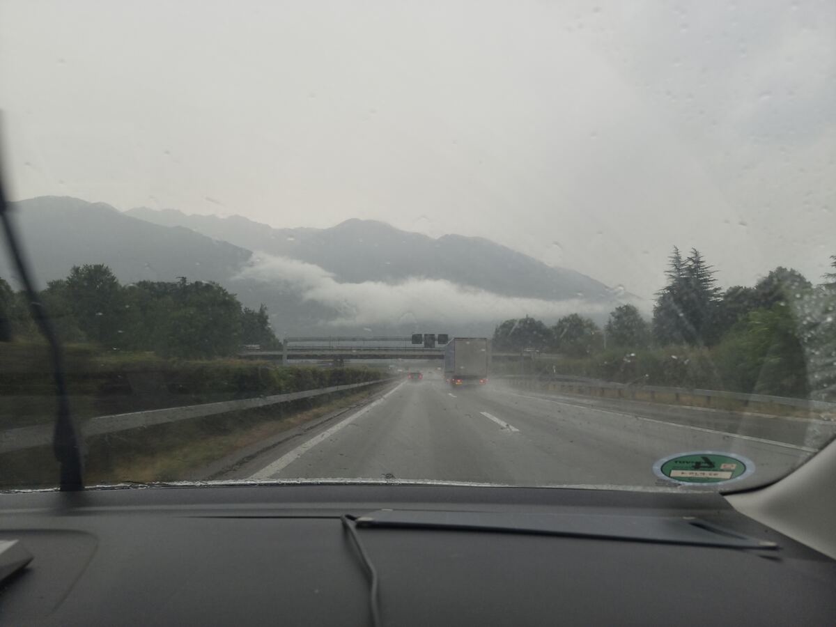 De foto is genomen vanuit de voorruit van een voertuig, met regendruppels duidelijk zichtbaar op het glas. Een ruitenwisser is gedeeltelijk zichtbaar aan de linkerzijde. Op het dashboard is een deel van de bovenkant te zien. Rechtsonder op de voorruit bevindt zich een groene ronde sticker met daarop 'TÜV' en het cijfer '02'. De weg is een natte, meerbaans snelweg met witte wegmarkeringen en een vangrail aan de linkerzijde. Enkele voertuigen, waaronder een vrachtwagen en personenauto's, rijden in