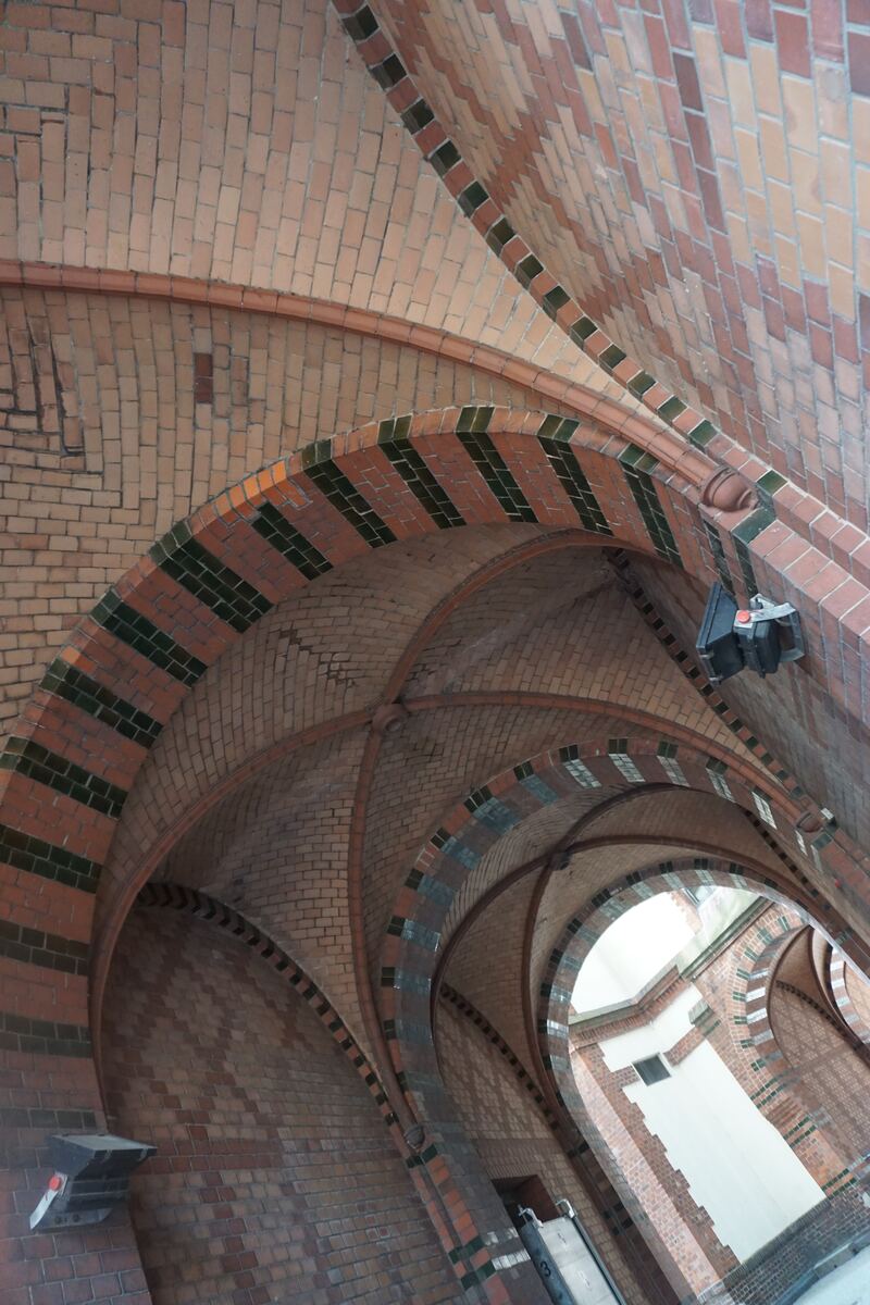De foto toont een interieur met meerdere bakstenen gewelven die een plafond vormen. De bakstenen zijn voornamelijk roodbruin van kleur en zijn in verschillende patronen gemetseld. Sommige bogen zijn versierd met stroken donkergroene of zwarte tegels. Er zijn structurele ribben zichtbaar die de gewelven ondersteunen. Aan een van de bogen hangt een modern ogend armatuur. Aan het einde van de gang is een helder verlichte opening te zien.
