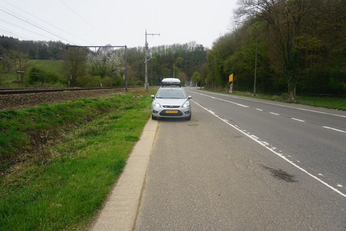 Een zilverkleurige Ford Focus stationwagen met een dakkoffer staat geparkeerd langs een tweebaansweg. Links van de auto liggen spoorrails en een groen talud. Op de achtergrond zijn beboste heuvels te zien onder een bewolkte lucht.