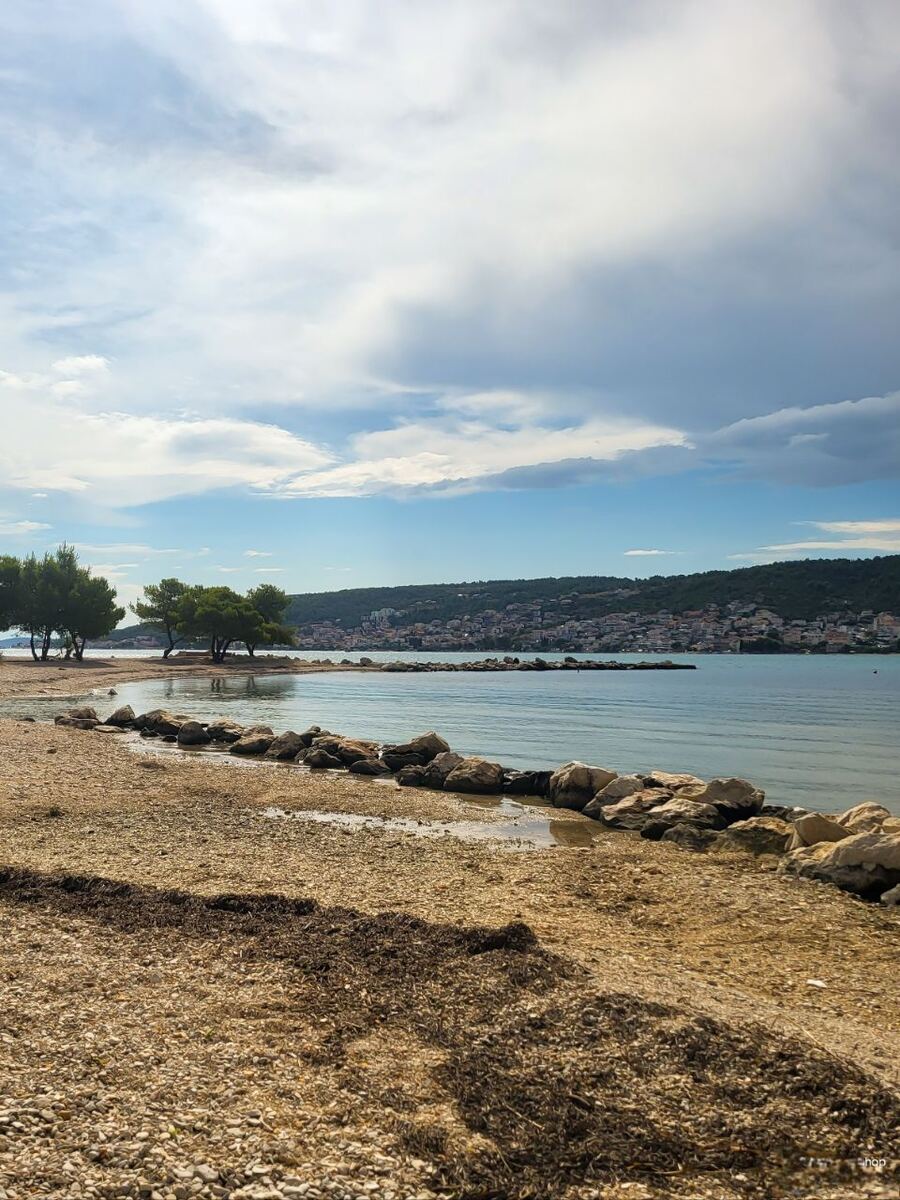 Een foto van een kiezelstrand met een strook zeewier op de voorgrond, langs een rustige baai. Aan de overkant van het water is een plaats met bebouwing zichtbaar, gelegen tegen een groene heuvel. Enkele bomen staan langs de kustlijn en de lucht is deels bewolkt.