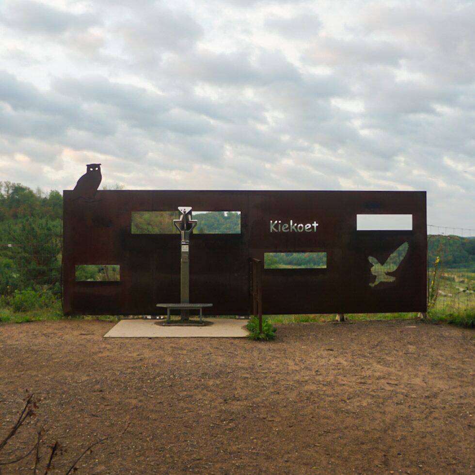 Een metalen informatiepaneel met uitgesneden figuren van een uil en een vogel, en het woord 'Kiekoot'. Ervoor staat een verrekijker op een paal. Achter het paneel is een groen landschap zichtbaar onder een bewolkte lucht.
