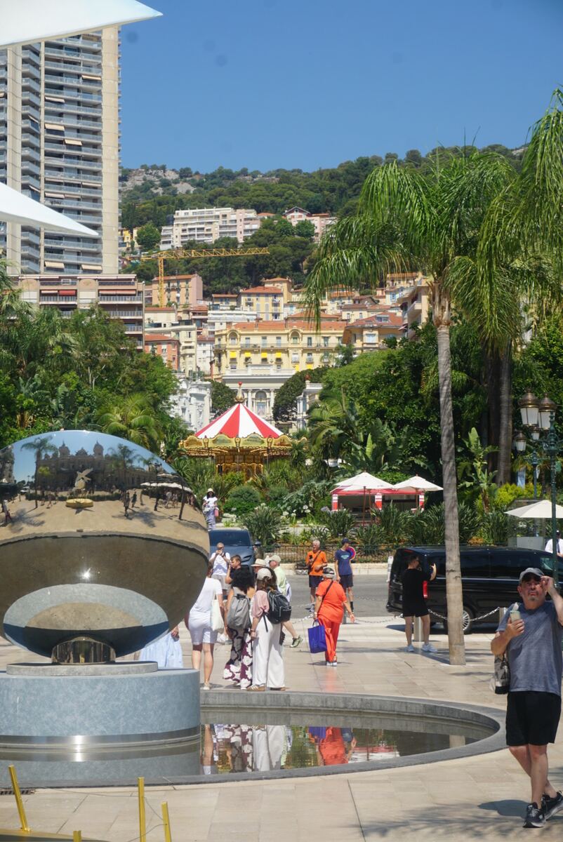 Een zonnige straat in Monaco met op de voorgrond een grote, reflecterende bol in een waterbassin. In de weerspiegeling zijn gebouwen, palmbomen en mensen te zien. Daarachter een park met een rood-wit gestreepte carrousel en diverse voetgangers. Op de achtergrond staan hellingen vol bebouwing en begroeiing onder een helderblauwe lucht.