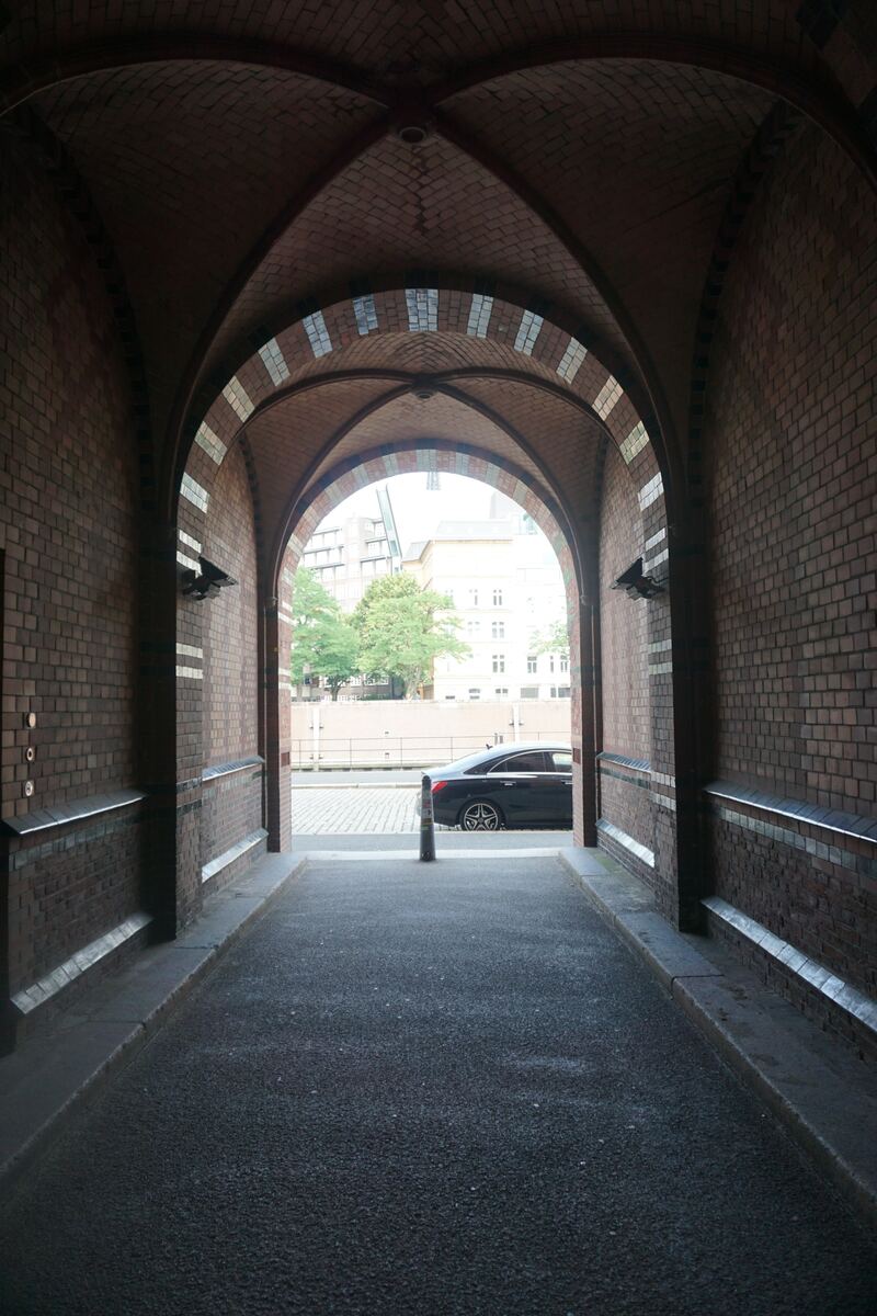 Een donkere, gewelfde doorgang van baksteen leidt naar een helderder verlichte straat. De boogconstructie heeft een plafond met decoratieve tegelpatronen en bakstenen muren met horizontale metalen of tegelbanden. Aan het einde van de doorgang is een geplaveide straat zichtbaar met een geparkeerde zwarte auto, een zwarte paal, een lage muur of hek, enkele bomen en gebouwen op de achtergrond. Het licht buiten is diffuus en bewolkt.