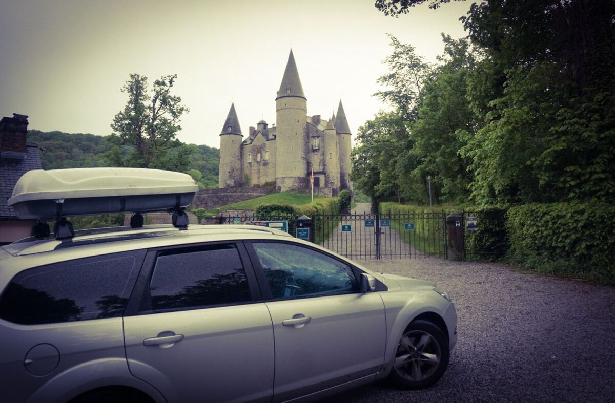 Een zilverkleurige Ford Focus stationwagen met een dakkoffer staat geparkeerd op een grindpad, met op de achtergrond het kasteel van Vêves. Het kasteel heeft meerdere kegelvormige torens en staat op een heuvel, omringd door bomen. Voor het kasteel is een metalen hek zichtbaar met bordjes en een haag.